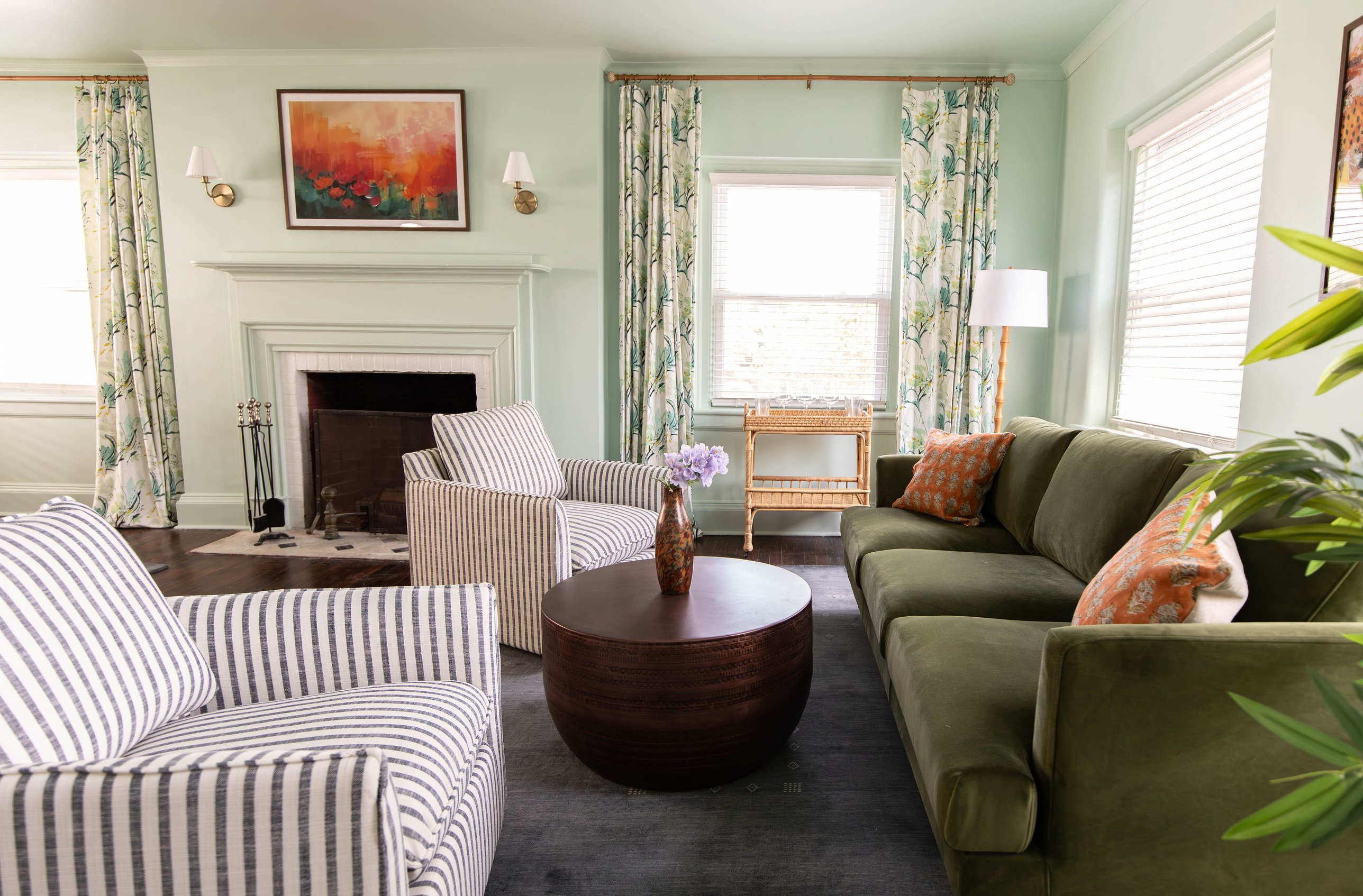 Mint-green living room with olive velvet sofa, striped swivel chairs, floral drapery and a painted brick fireplace by K&B Interiors